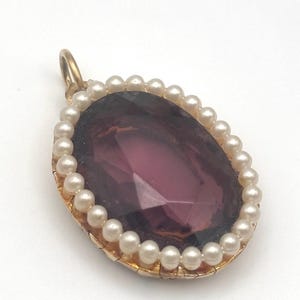 May include: A gold-toned pendant with a large, faceted, oval-shaped, deep purple stone. The stone is surrounded by a border of small, white pearls. The pendant has a small loop at the top for a chain.