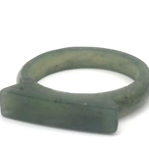 May include: A smooth, translucent sage green ring featuring a flat, rectangular bar element integrated into the circular band. The material displays subtle natural variations in color and texture throughout the entire surface of the ring.