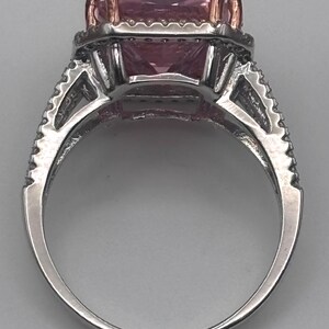 May include: A silver ring with a large, square, pink gemstone. The ring has a halo of smaller stones around the main gem and additional detailing on the band. The prongs holding the gemstone are gold-colored.