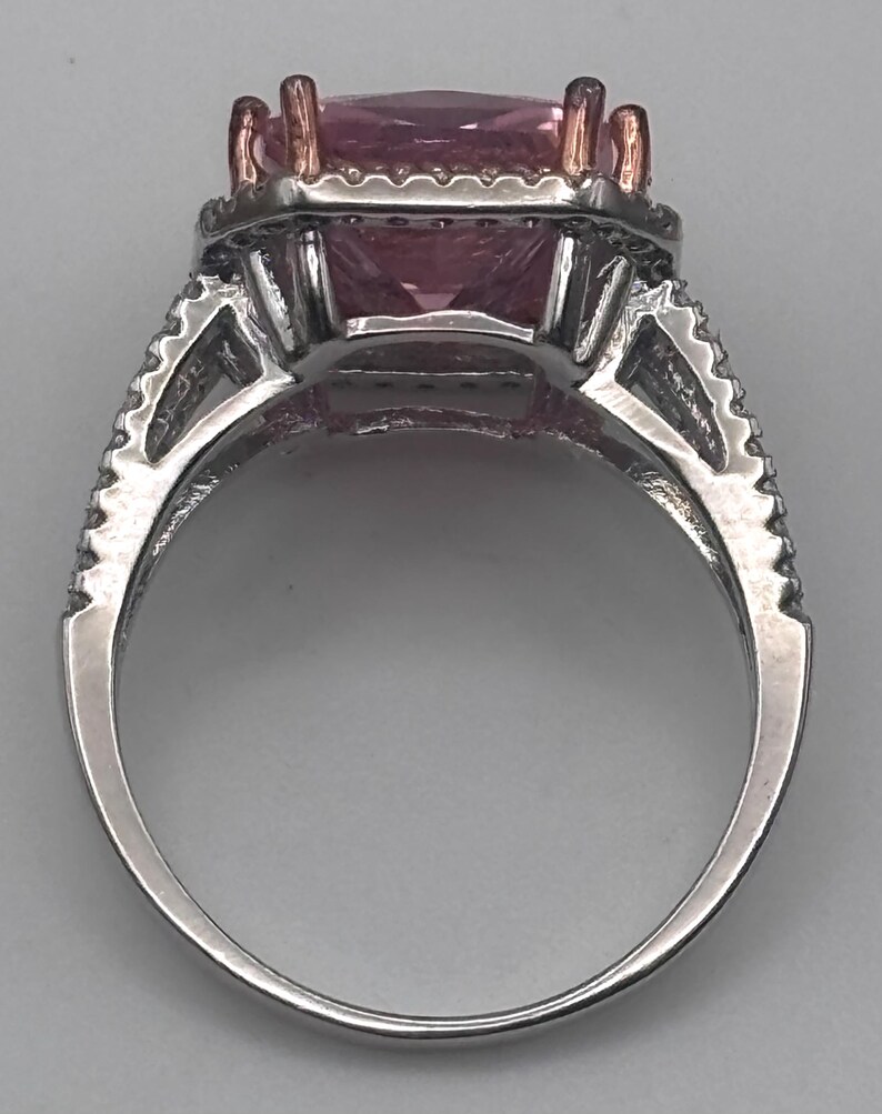May include: A silver ring with a large, square, pink gemstone. The ring has a halo of smaller stones around the main gem and additional detailing on the band. The prongs holding the gemstone are gold-colored.