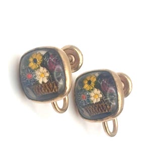 May include: A pair of vintage clip-on earrings. Each earring features a square, gold-toned frame encasing a miniature floral arrangement. The arrangement includes yellow, white, and blue flowers in a brown basket against a dark blue background.