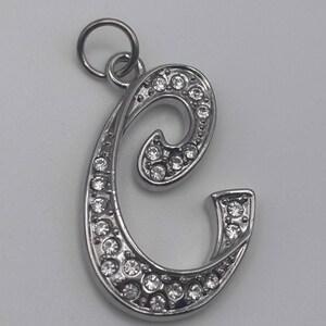 May include: Silver-toned pendant in the shape of the letter "C", encrusted with numerous small, clear crystals. The pendant has a loop at the top for a chain. The crystals are set into the metal, creating a sparkling effect. This jewelry item is suitable for necklaces or bracelets.