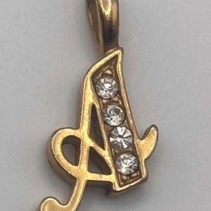May include: Gold-toned pendant in the shape of the letter "A", embellished with four clear, round gemstones. The pendant has a loop at the top for a chain and a decorative flourish at the bottom.