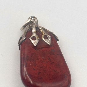 May include: A red pendant with a silver-colored bail. The pendant is a smooth, oblong stone with dark red and brown marbling. The top of the stone is capped with ornate silver-colored metalwork.