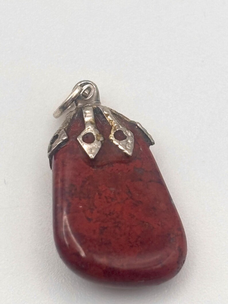 May include: A red pendant with a silver-colored bail. The pendant is a smooth, oblong stone with dark red and brown marbling. The top of the stone is capped with ornate silver-colored metalwork.