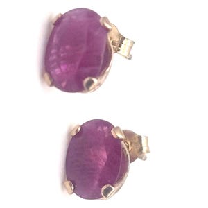 May include: A pair of stud earrings featuring oval-shaped, deep magenta gemstones set in gold-colored metal. The earrings have a post and butterfly backing. The gemstones have a polished, reflective surface.