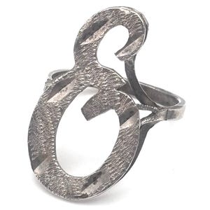 May include: A silver-toned ring featuring an open, stylized letter "S" design. The ring has a textured surface with diagonal etched lines. The band is simple and connects to the base of the letter.