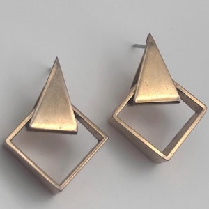 May include: A pair of gold-toned metal stud earrings featuring a geometric design. Each earring consists of a solid triangle shape positioned over an open square frame, creating a layered, angular appearance with a metallic finish and post backs.