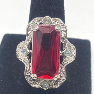 May include: A silver ring with a large, red, rectangular gemstone surrounded by smaller, iridescent stones.
