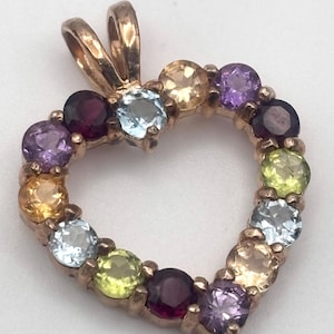 May include: A heart-shaped pendant with a gold-colored frame, adorned with a variety of colorful gemstones. The gemstones include shades of purple, yellow, blue, and green. The pendant has a loop at the top for a chain.