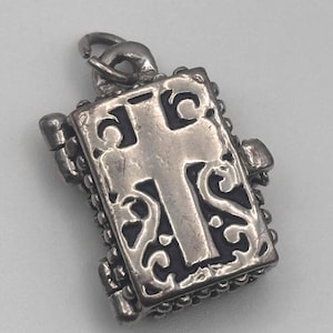 May include: A silver-toned pendant with a cross design. The pendant has an ornate, openwork border and a bail for attaching to a chain. The cross is centered on the pendant and is also silver-toned.