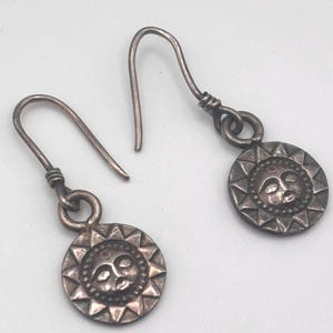 May include: A pair of silver-toned earrings. Each earring features a circular pendant with a sun face design at the center, surrounded by triangular rays. The earrings have a hook closure.
