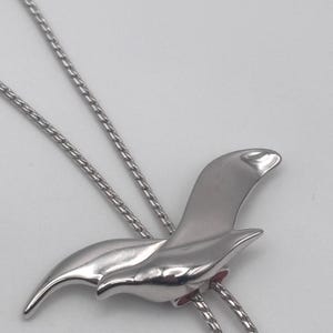 May include: A silver-toned necklace featuring a stylized bird pendant. The pendant has a smooth, polished finish and a curved, wing-like design. The necklace chain is a delicate, linked style. The bird's body has a small, red accent.