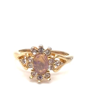 May include: A gold-toned ring featuring a central oval-shaped, light pink gemstone surrounded by clear, round accent stones. The ring's band has a decorative design on each side, creating a floral-like appearance. The ring is set against a white background.