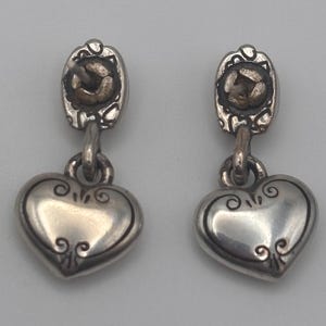 Op de afbeelding: Een paar zilverkleurige oorhangers. Elke oorbel heeft een hartvormige bedel met decoratieve krullen en een kleine, ovaalvormige bovenkant met een getextureerd midden. De oorbellen zijn verbonden door een kleine lus.