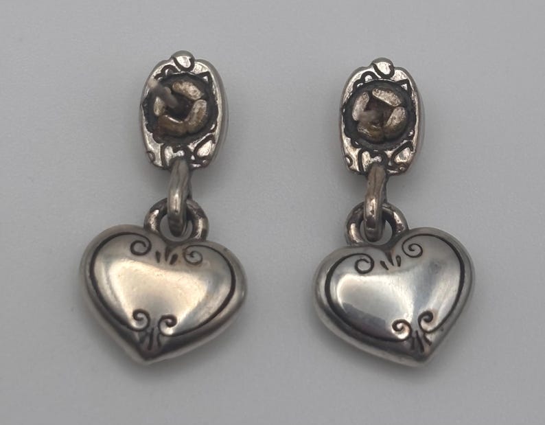 Op de afbeelding: Een paar zilverkleurige oorhangers. Elke oorbel heeft een hartvormige bedel met decoratieve krullen en een kleine, ovaalvormige bovenkant met een getextureerd midden. De oorbellen zijn verbonden door een kleine lus.