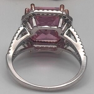 May include: A silver ring with a large, rectangular pink gemstone as the centerpiece. The gemstone is surrounded by smaller, clear stones and set in a detailed silver band. The ring has a classic, elegant design.