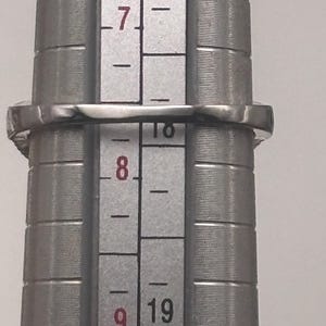 May include: A silver ring with a square gemstone is being measured on a ring sizer. The ring is placed on the sizer between sizes 7 and 8. The ring is made of a shiny, reflective metal.