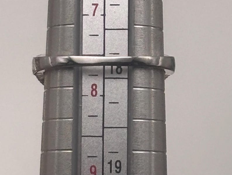May include: A silver ring with a square gemstone is being measured on a ring sizer. The ring is placed on the sizer between sizes 7 and 8. The ring is made of a shiny, reflective metal.