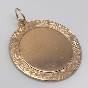 May include: A gold-colored pendant with an oval center and a decorative wave pattern around the edge. The pendant has a small loop at the top for a chain or cord. The overall design is simple and elegant.