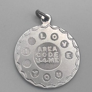 May include: A silver-colored pendant with the words "AREA CODE GAME" in the center. The pendant has a scalloped edge and the words "I LOVE YOU" are arranged around the center. The pendant has a small loop at the top for a chain.