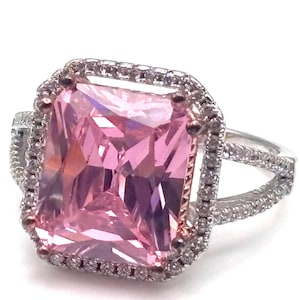May include: A silver ring featuring a large, rectangular pink gemstone surrounded by a halo of smaller, clear stones. The ring has a split shank design, adding to its elegant appearance. The setting is detailed and eye-catching.