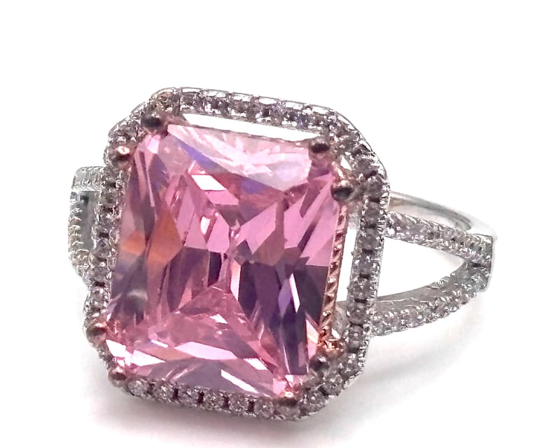 May include: A silver ring featuring a large, rectangular pink gemstone surrounded by a halo of smaller, clear stones. The ring has a split shank design, adding to its elegant appearance. The setting is detailed and eye-catching.