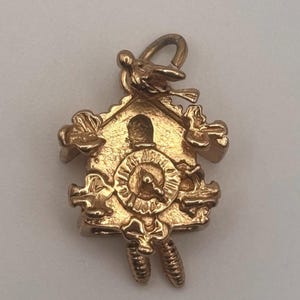 May include: A gold-colored charm in the shape of a cuckoo clock. The charm features a bird perched on top, floral accents, and a clock face with Roman numerals. The pendulum is visible at the bottom. The charm is likely made of metal.
