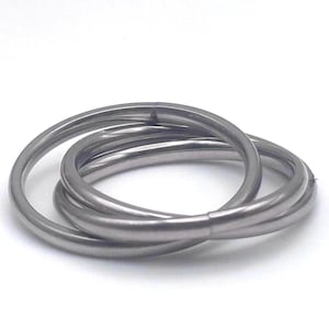 May include: A set of three interlocking silver-toned metal rings with a smooth, polished finish. The rings are connected in a continuous loop design, creating a layered appearance with a consistent circular shape and a reflective metallic surface.