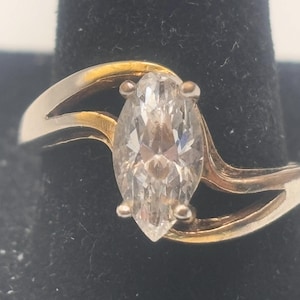 May include: A gold ring with a large, clear marquise-shaped gemstone. The ring band is a double band with a wave-like design.
