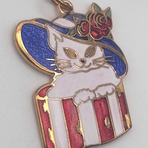 May include: A gold-toned cat pendant with a blue and white hat adorned with red roses. The cat is white with gold eyes and whiskers, and the base is red and white striped. A charming, vintage-style cat charm.