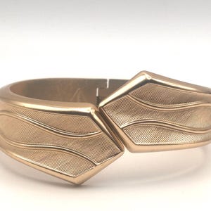 May include: A gold-toned hinged bangle bracelet with a wave-like pattern. The bracelet is made of metal and has a polished finish.