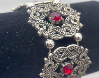 May include: Silver-toned bracelet with ornate filigree design and three vibrant red gemstones. The bracelet is composed of two main sections connected by smaller, beaded links. The intricate metalwork features swirling patterns and small, decorative details.