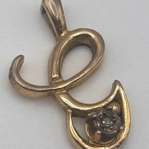 May include: Gold-toned pendant in the shape of the letter "L", featuring a small, round, gray gemstone. The pendant has a loop at the top for a chain. The design is ornate and detailed, with a vintage aesthetic.