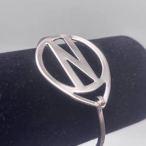 May include: Silver bracelet with a circle design featuring the letter N cut out. The bracelet is open and has a simple hinge.