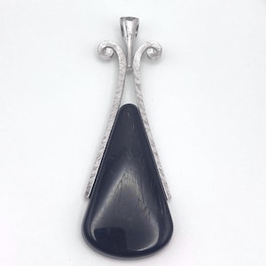 May include: A silver-toned pendant with a teardrop-shaped, dark gray stone. The pendant has a decorative, textured metal frame with a loop at the top for a chain. The stone has a smooth, polished surface.
