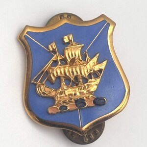 May include: A shield-shaped lapel pin featuring a gold-toned sailing ship centered on a smooth, light blue enamel background. The pin has a raised gold border and a metallic finish, with a small, circular fastener visible on the back side.