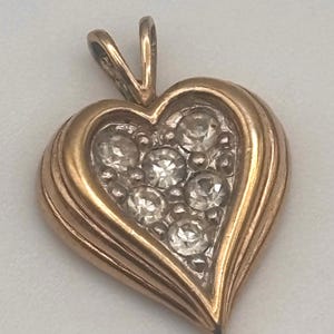 May include: A heart-shaped pendant crafted from a warm-toned metal, featuring a tiered, ridged border. The center of the heart is set with seven small, clear, round-cut stones arranged in a cluster, and it includes a triangular bail at the top.