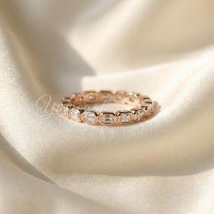 May include: Rose gold eternity ring featuring emerald-cut gemstones. The ring is set with multiple rectangular-shaped stones, creating a continuous band. The ring is displayed on a white satin surface, highlighting the ring's sparkle and design.