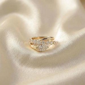 May include: Two gold rings with clear gemstones. One ring features a pear-shaped stone, while the other has a row of smaller, round stones. The rings are set against a cream-colored, silky background.