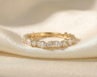 May include: A gold ring with a continuous band of alternating round and marquise-cut clear gemstones. The ring is set on a cream-colored fabric, showcasing its sparkle and design. The ring is a piece of jewelry.