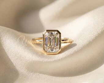 May include: A gold ring with an emerald-cut diamond. The diamond is set in a bezel setting and has a rectangular shape. The ring is on a white satin background.