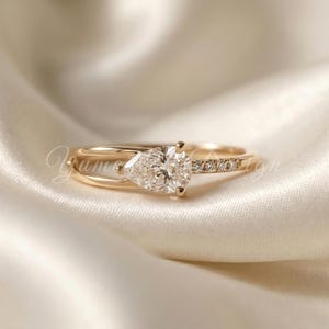 May include: A gold engagement ring with a pear-shaped diamond center stone. The band features a split shank design and small accent diamonds. The ring is set against a soft, white background.