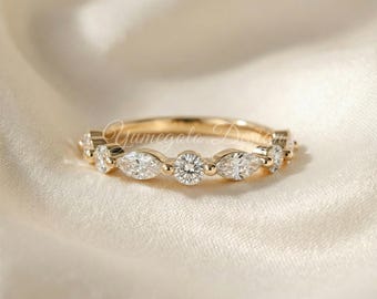 May include: A gold band ring with alternating round and marquise-cut clear gemstones. The ring is set on a cream-colored fabric. The ring has the text "Yumegoto Design" engraved on the side.
