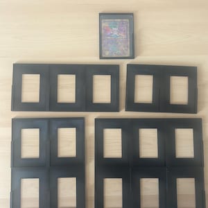 May include: Black card display frames with rectangular openings are arranged on a light wood surface. A single card is displayed in a black frame. The card has a holographic design with various colors. The frames are designed to hold multiple cards.
