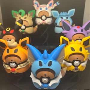 May include: A collection of eight small, round, 3D-printed figurines, each featuring a brown bear-like character wearing a different colored hood inspired by various creatures. The figures are arranged in a circular display, showcasing vibrant colors including blue, yellow, orange, black, purple, and pink, with each character holding a small circular emblem on its chest.