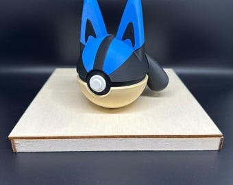 Könnte beinhalten: Eine blaue und schwarze Pokémon-Figur in Form eines Pokéballs mit einem weißen Knopf auf der Vorderseite. Die Figur steht auf einem Holzsockel.