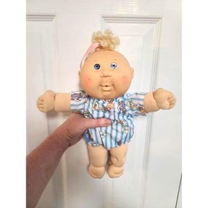 May include: A vintage Cabbage Patch Kid doll with a bald head, blue eyes, and rosy cheeks. The doll wears a blue and white striped onesie with a pattern of teddy bears and other toys. A small tuft of blonde hair is tied with a pink ribbon.