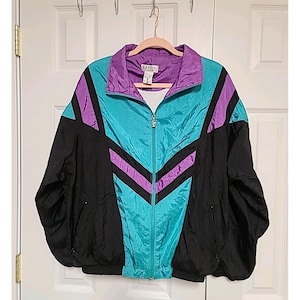 May include: A vintage Lavon jacket in a vibrant color block design. The jacket features a teal, purple, and black color scheme with a zip-up front and a collared neckline. The jacket is hanging on a wooden hanger.