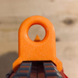 May include: Close-up of an orange sight on a black and red object. The sight has a circular opening and a triangular aiming point. The background is a wooden surface.